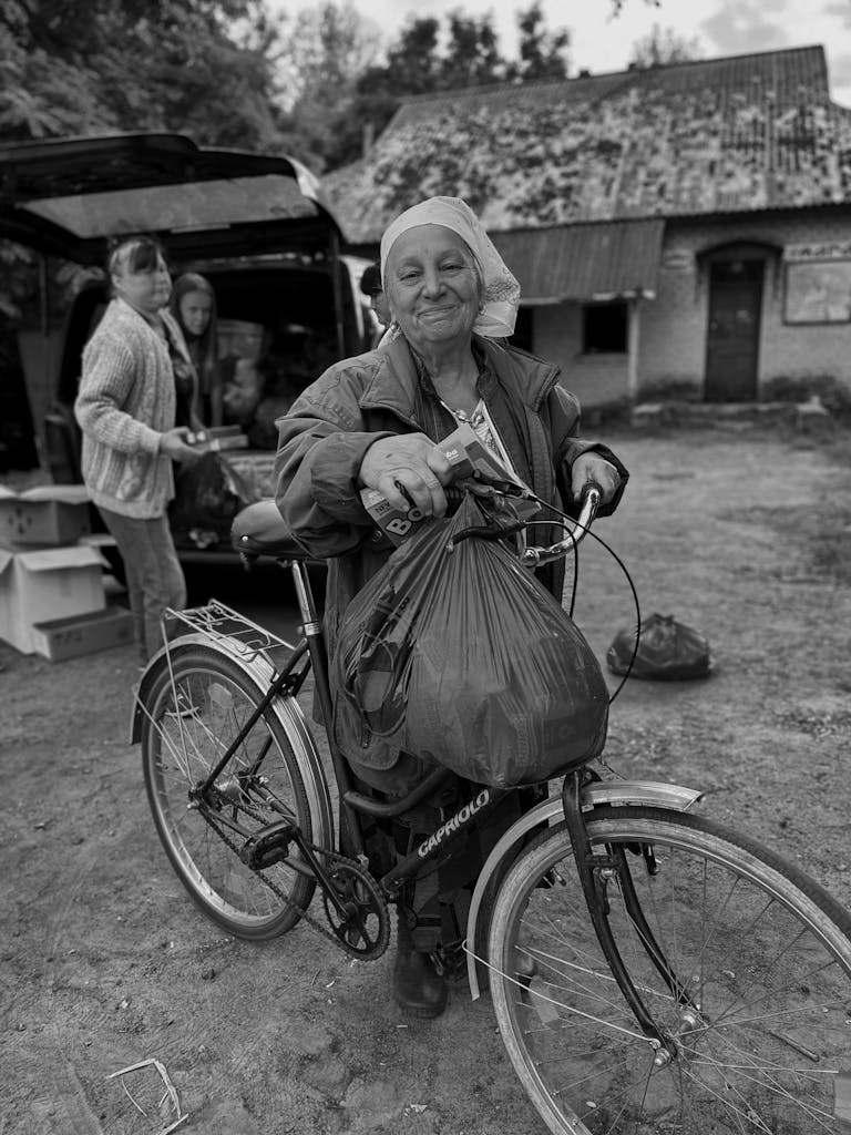 Elderly woman smiling while holding a bicycle filled with groceries in a Ukrainian village.